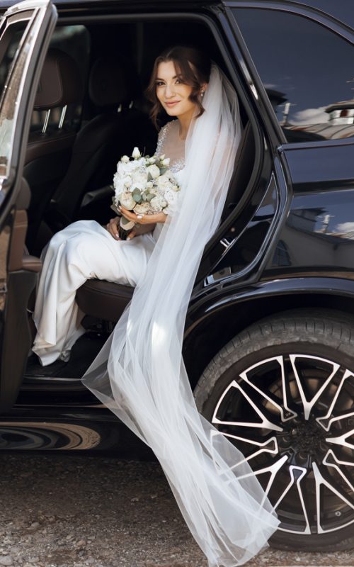 Jeune mariée en robe blanche et voile sortant d'une voiture de luxe avec chauffeur VTC pour son mariage.